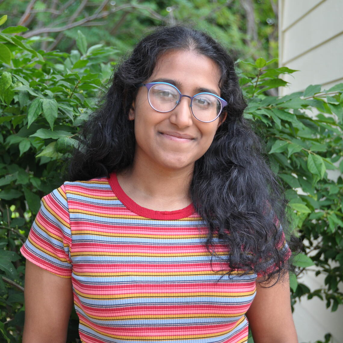 Tarini Fernando smiles outside in front of a bush. She is wearing a striped t-shirt and glasses.