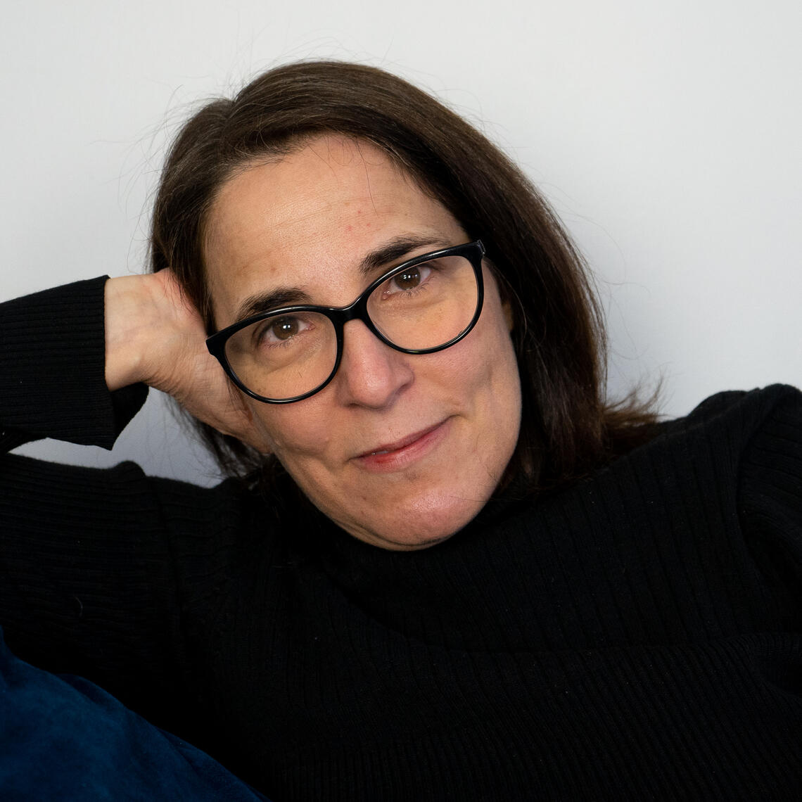Xenia Reloba de la Cruz leans on one arm and smiles in front of a white background. She is wearing a black turtleneck and black glasses.